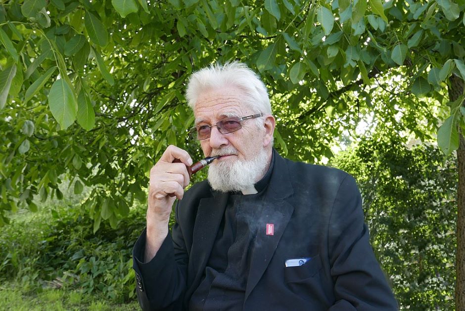 Starszy mężczyzna z brodą pali fajkę w otoczeniu zieleni.
