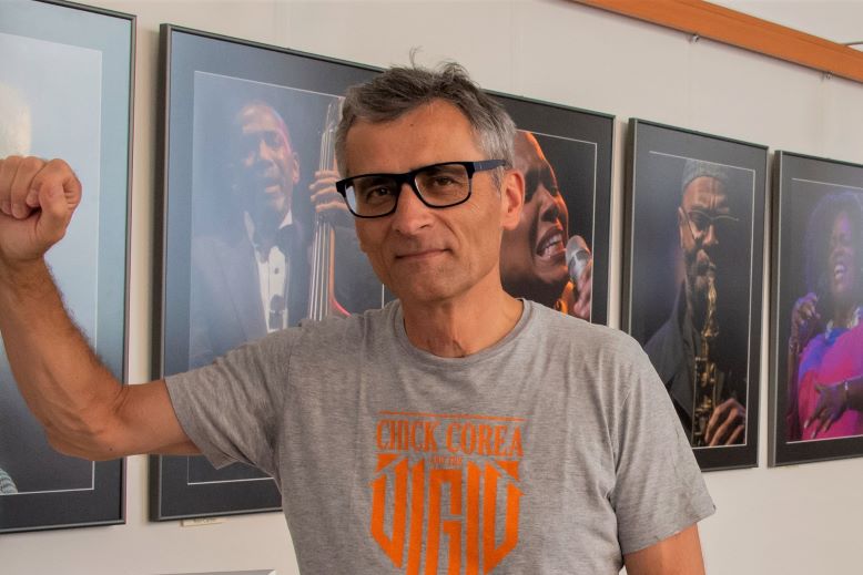 Mężczyzna w okularach na tle wiszących na ścianie zdjęć muzykó jazzowych.