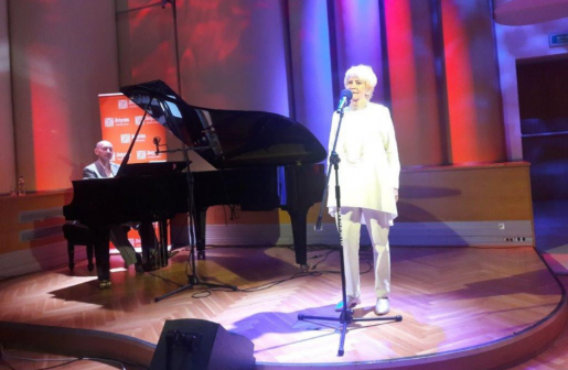 Na scenie kobieta przy mikrofonie, po lewej stronie mężczyzna przy fortepianie.