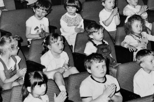 Czarno-białe zdjęcie. Widownia pełna siedzących kilkuletnich dzieci. Atmosfera zainteresowania.