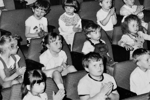 Czarno-białe zdjęcie. Widownia pełna siedzących kilkuletnich dzieci. Atmosfera zainteresowania.