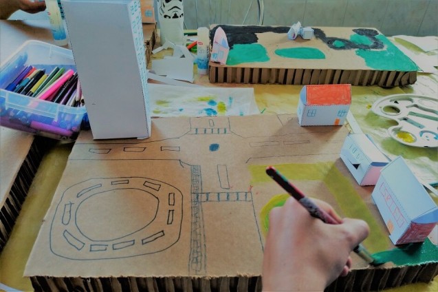 Stół, na którym stowi kartonowa makieta miasta. Dziecięca ręka rysuje ulice.